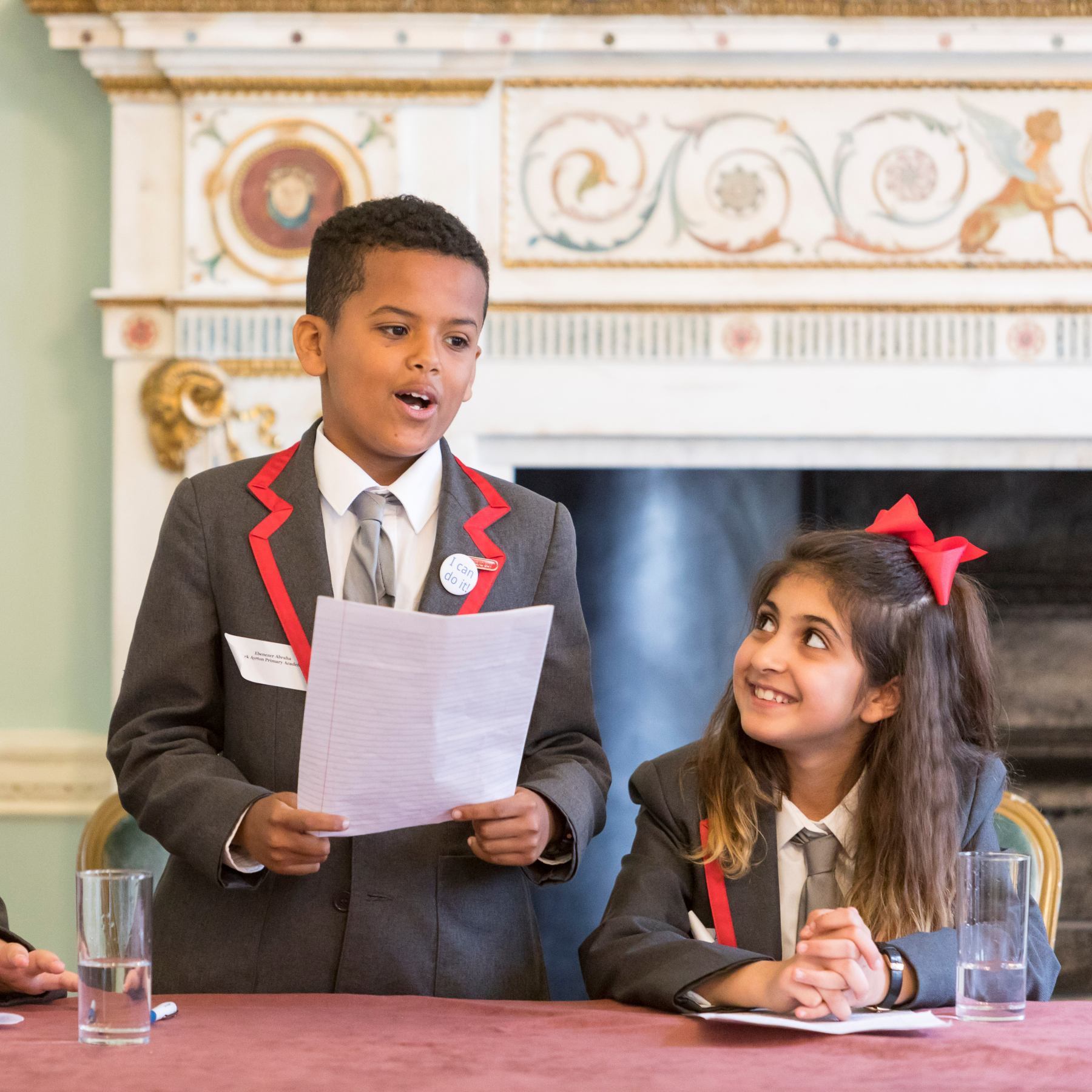 Two primary school students (male and female). The male standing with a piece of paper in his hand and reading from it. The female sitting down and smiling at the her colleague.