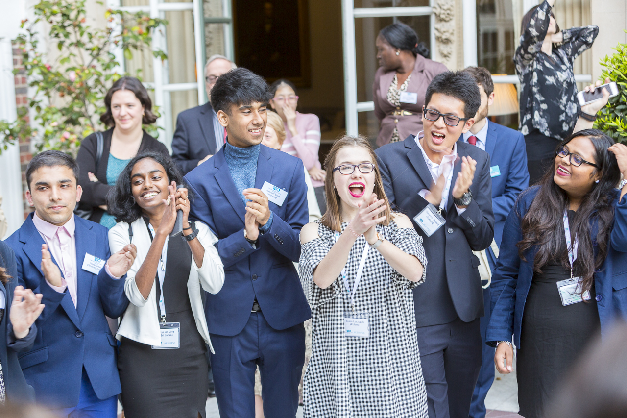 A group of secondary school students standing outside a building while clapping and cheering.
