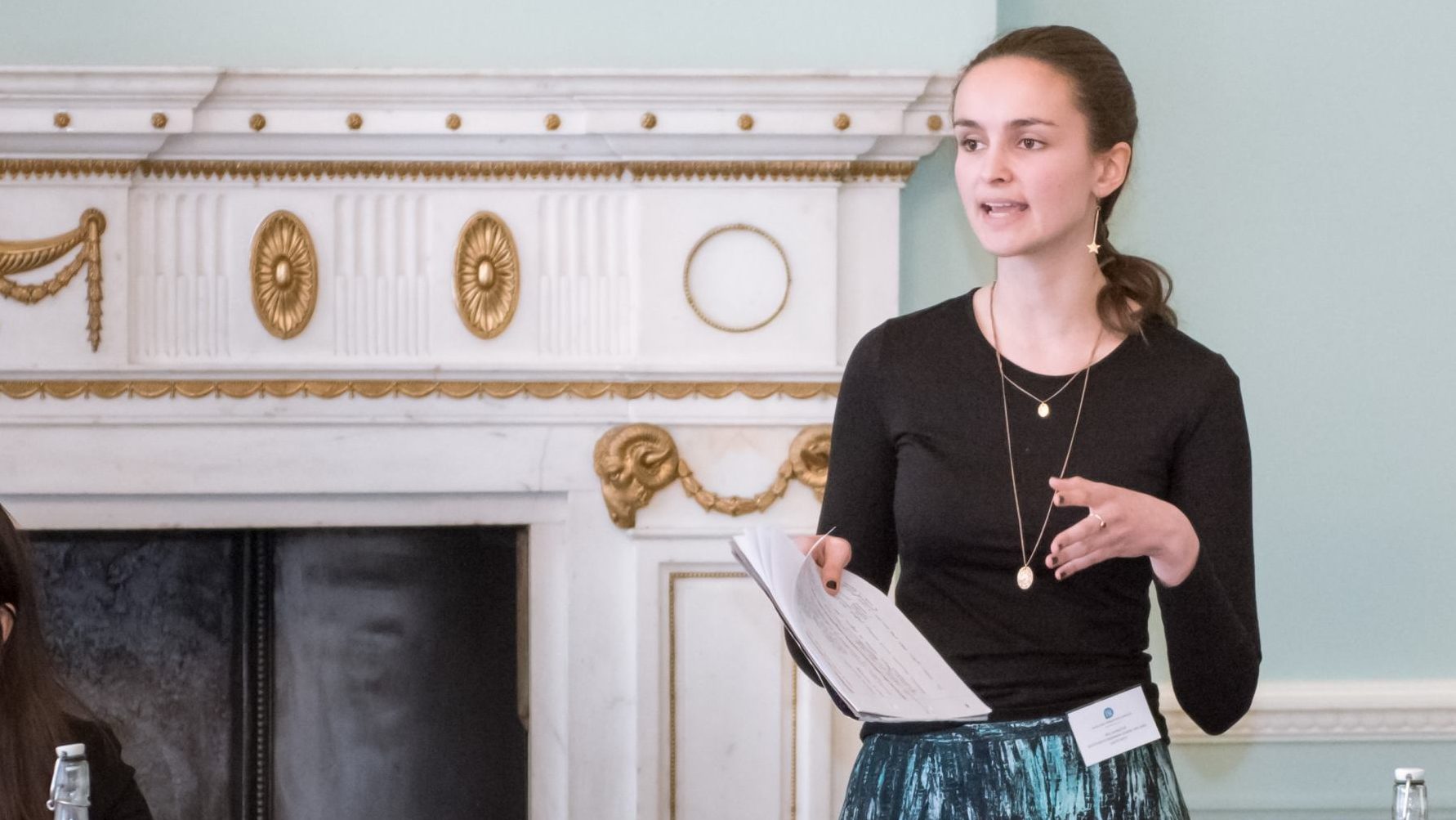 A picture of a young female debater standing in front of a fireplace inside the ESU London's office building at Dartmouth House.